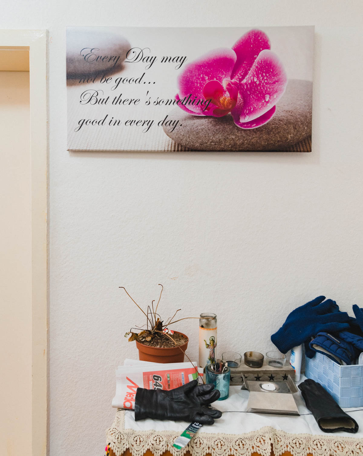 Die Wand einer Wohnung: Ein Bild einer pinken Blüte, verziert mit einer Lebensweisheit, darunter eine Ablage, auf der unter anderem Handschuhe, Teelichter und ein Werbeprospekt liegen Die Wand einer Wohnung: Ein Bild einer pinken Blüte, verziert mit einer Lebensweisheit, darunter eine Ablage, auf der unter anderem Handschuhe, Teelichter und ein Werbeprospekt liegen (Foto: Christian Protte)