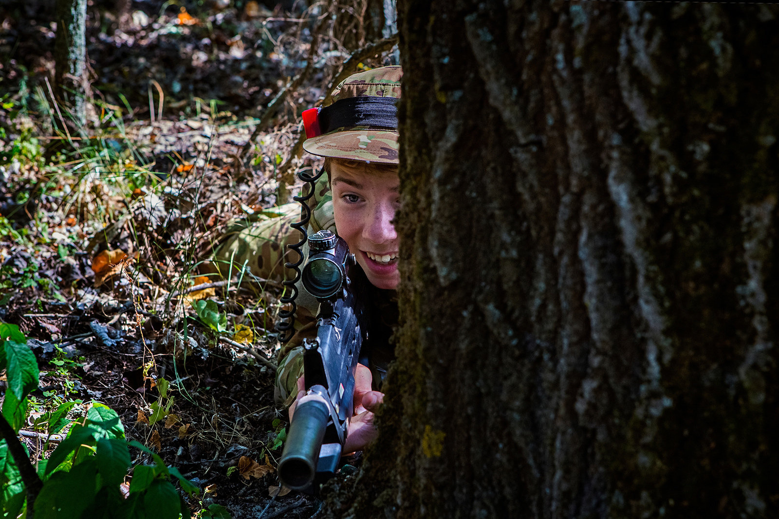 Ein Junge lauert mit einem Gewehr hinter einem Baum 