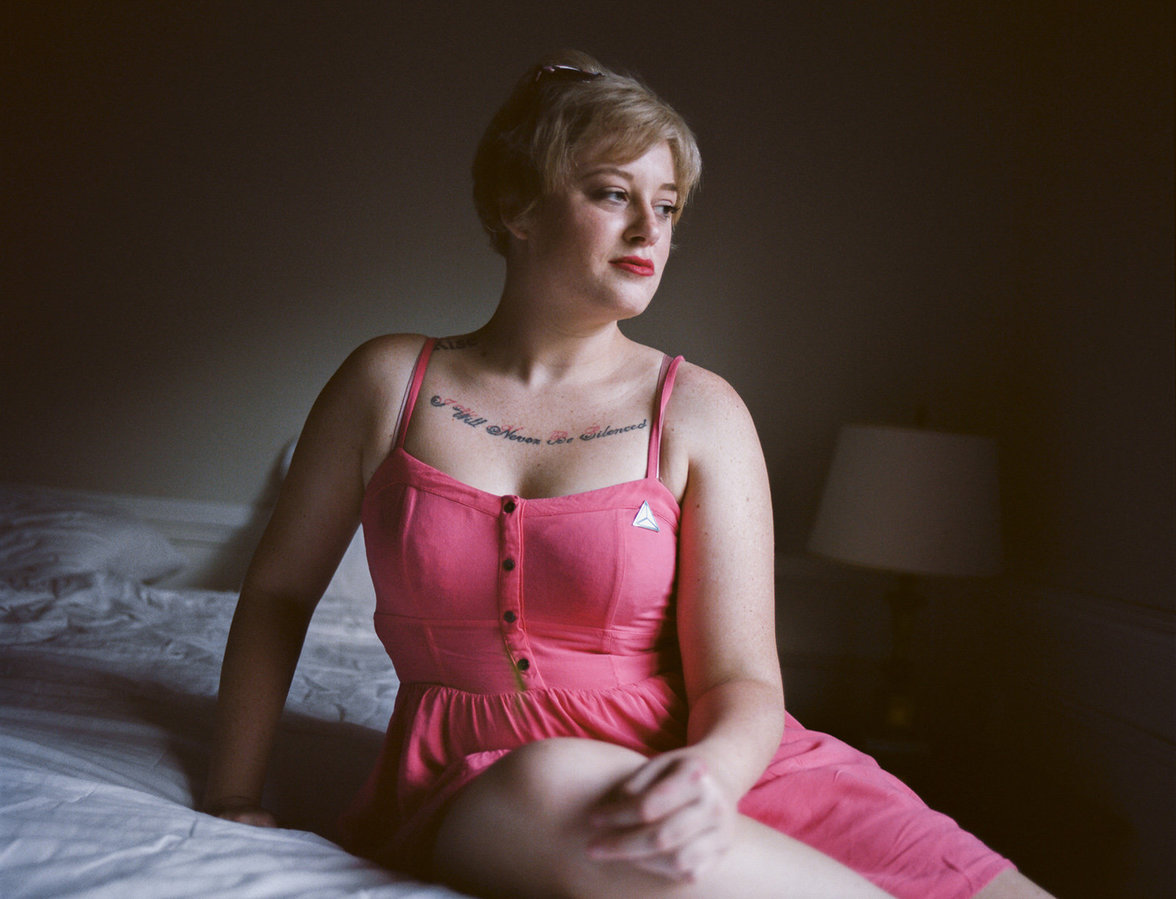 Erika sitzt in einem rosa Kleid auf einem Bett, ihr Tattoo "I will never be silenced" ist zu sehen
