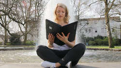 Eine Frau sitzt vor einem Springbrunnen und liest in einem aufgeklappten Laptop, als wäre es ein Buch