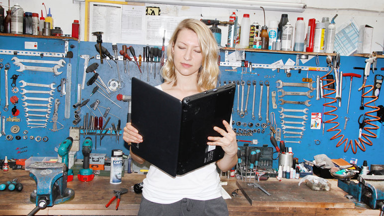 Eine Frau steht in einer Werkstatt und liest in einem aufgeklappten Laptop wie in einem Buch. 