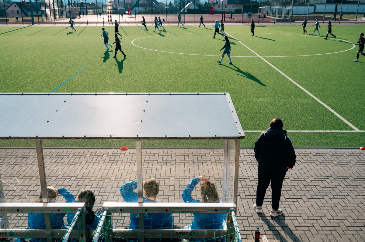 Kian (rechts) und ihre Mitspielerinnen sitzen beim Spiel auf der Ersatzbank und schauen aufs Spielfeld
