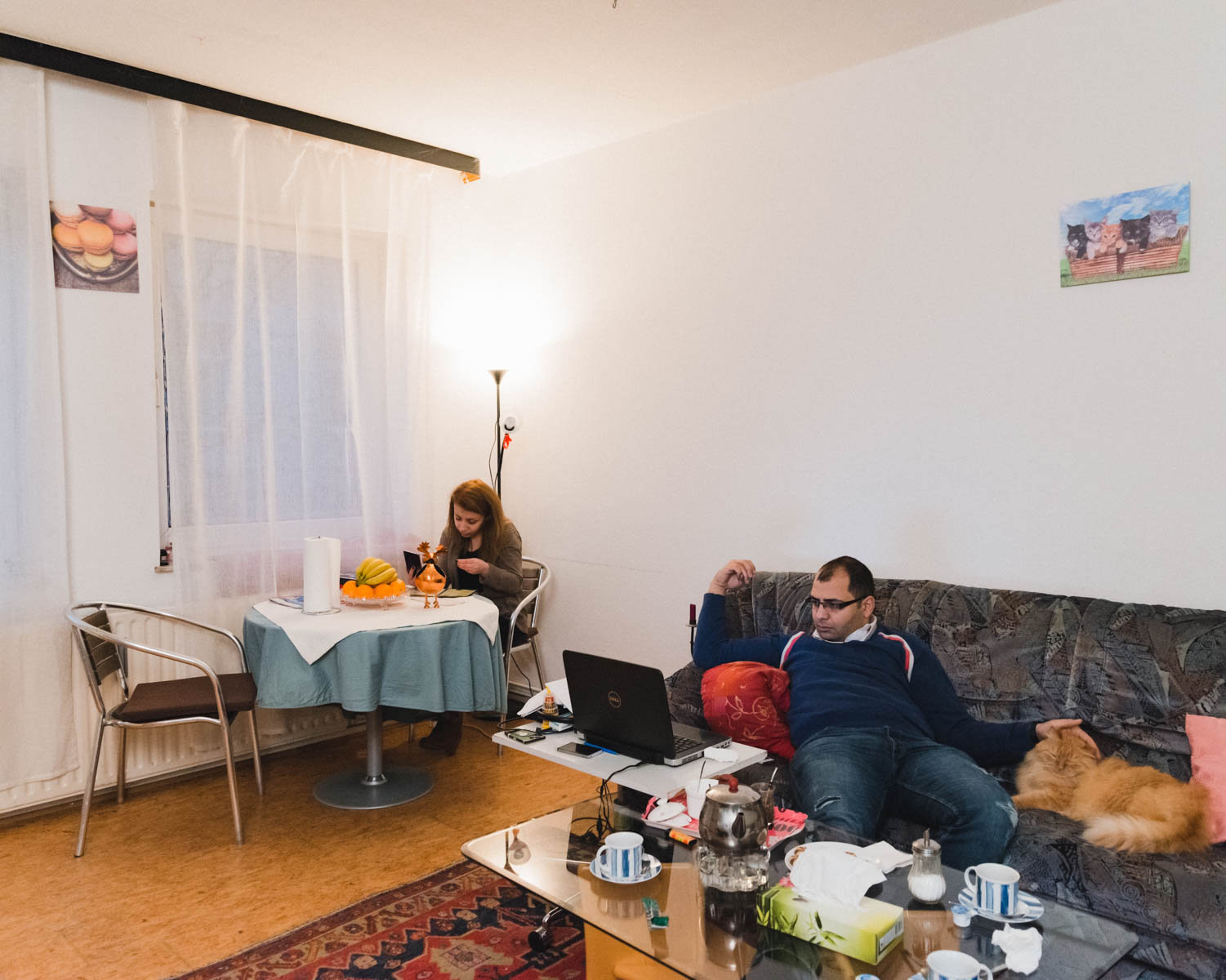 Ein Ehepaar in seinem Wohnzimmer: Aziz lümmelt mit dem Laptop auf der Couch, neben ihm liegt ein Kater. Lama sitzt am Tisch (Foto: Christian Protte)