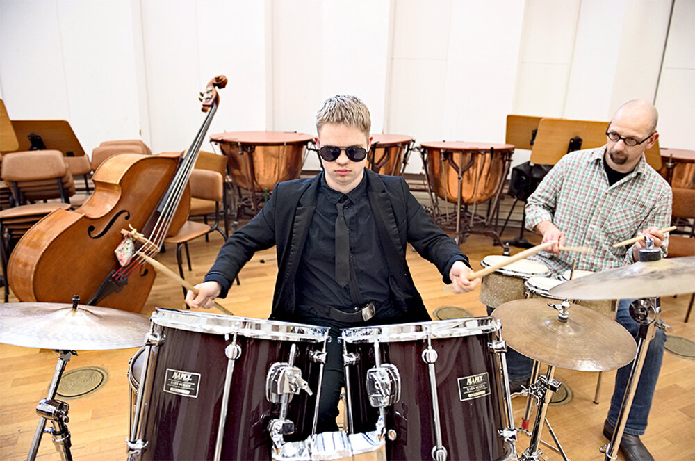 Ein junger Mann mit Sonnenbrille und schwarzem Jackett sitzt an einem Schlagzeug