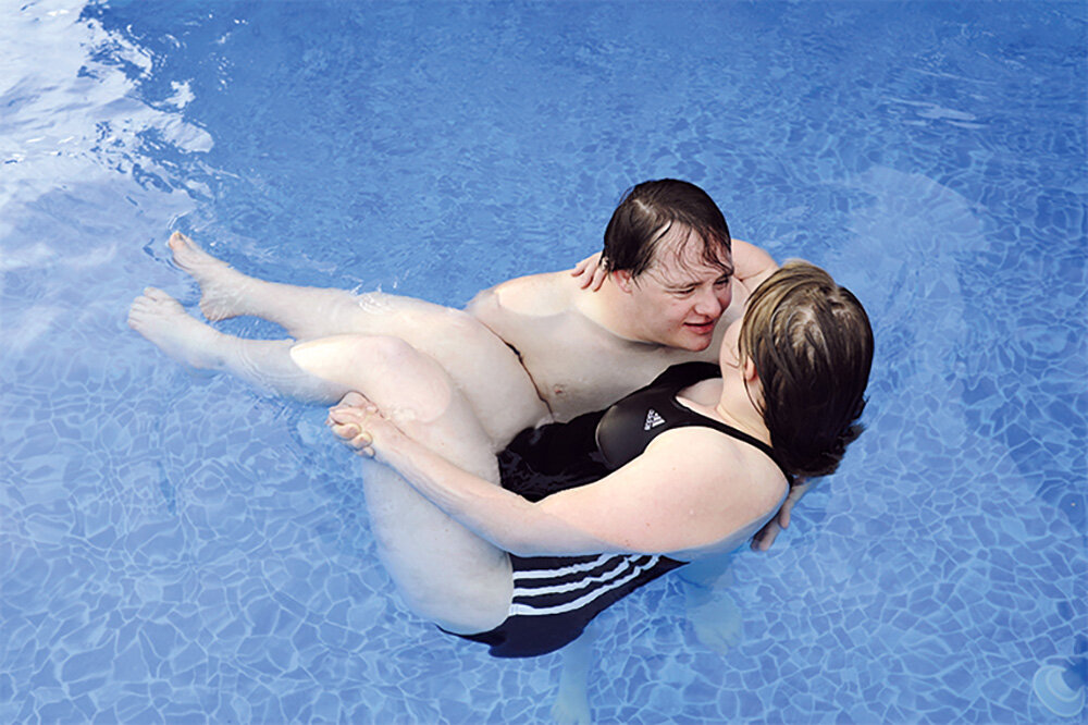 Ein Mann trägt eine Frau, beide befinden sich in einem Schwimmbecken